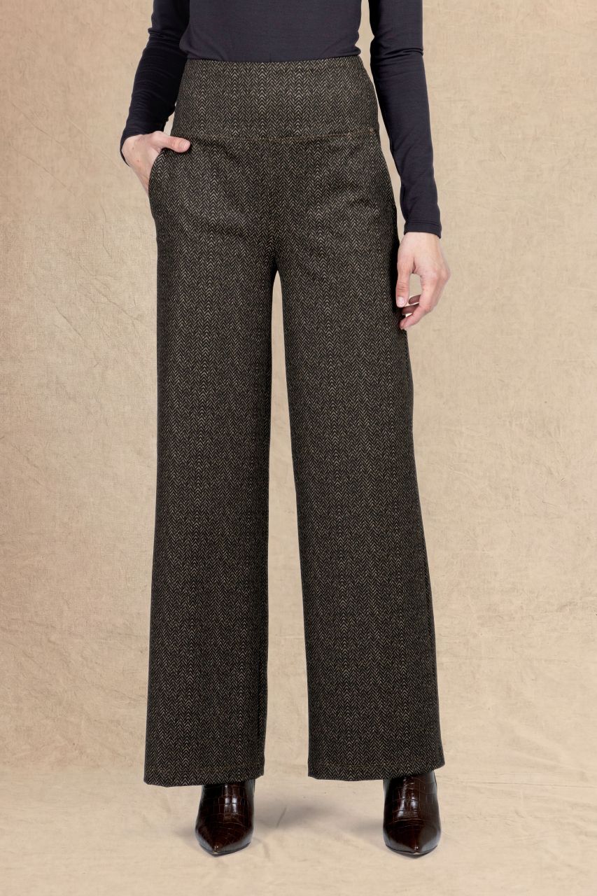 A model wearing high-waisted, wide-leg trousers in a dark brown and black herringbone pattern, paired with a dark long-sleeve top and dark brown pointed-toe boots, set against a textured beige backdrop.