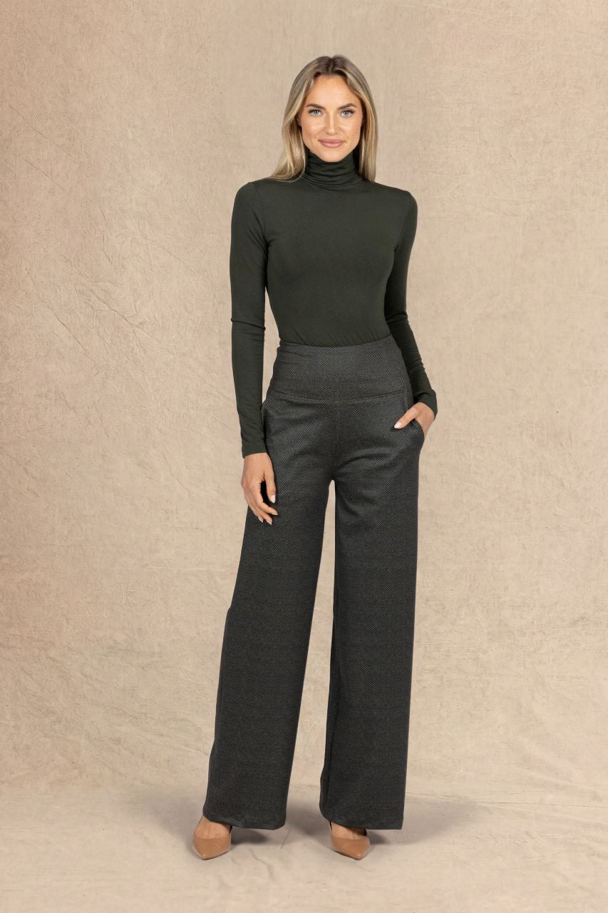 A woman modeling a dark loden green ribbed turtleneck sweater and dark olive tweed wide-leg trousers, standing against a textured beige studio backdrop.