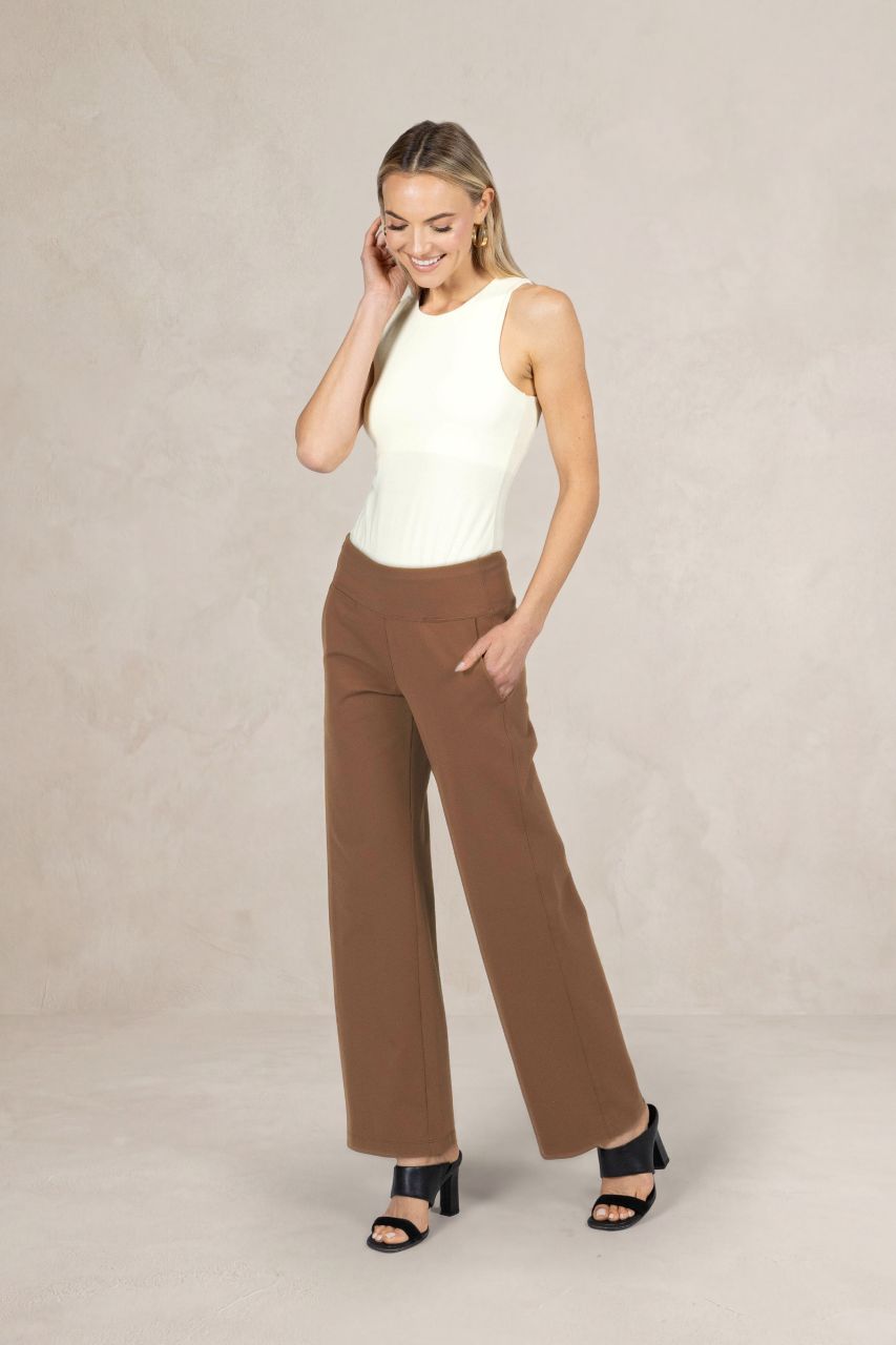 Woman wearing a white sleeveless tank top and brown mid-rise pants on a plain background