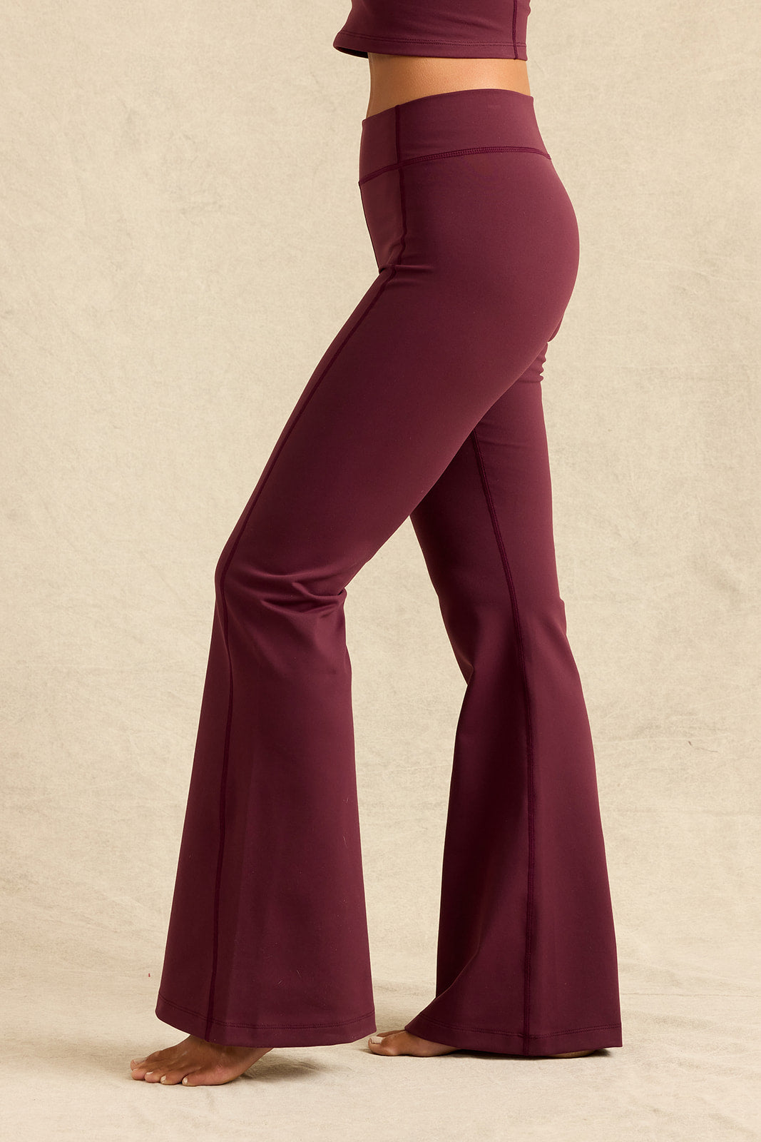 A person wearing high-waisted, wide-leg flared pants in a deep burgundy shade, standing barefoot against a plain beige studio backdrop.