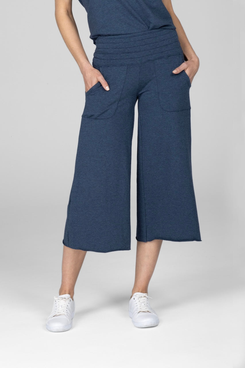 Front view of model wearing a pair of heather lake blue cropped Boho yoga pants with a smocked waistband and hands in front pockets, worn with white sneakers.