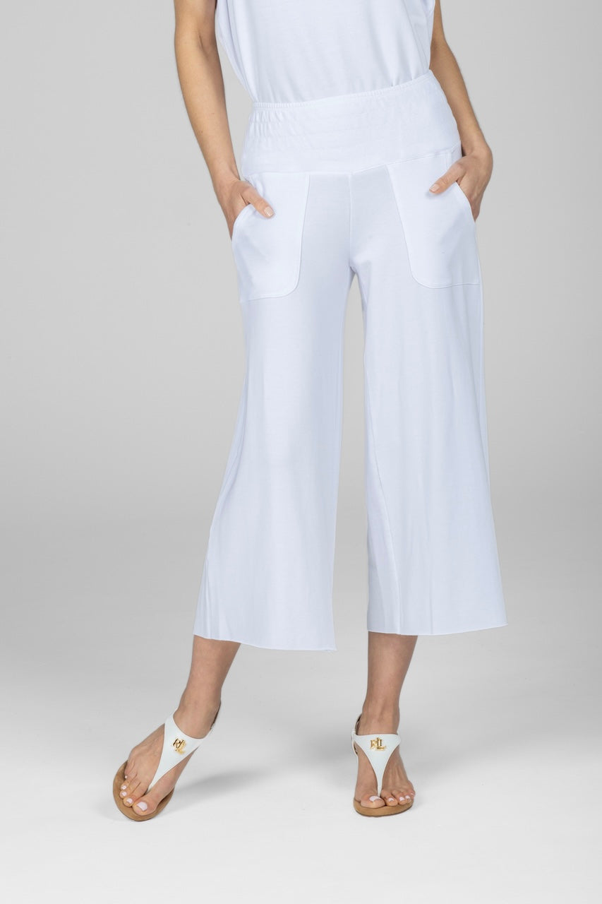 Front view of a woman modeling a pair of white cropped Boho yoga pants with a smocked waistband and large pockets, paired with a white top and white sandals with hands in both front pockets.