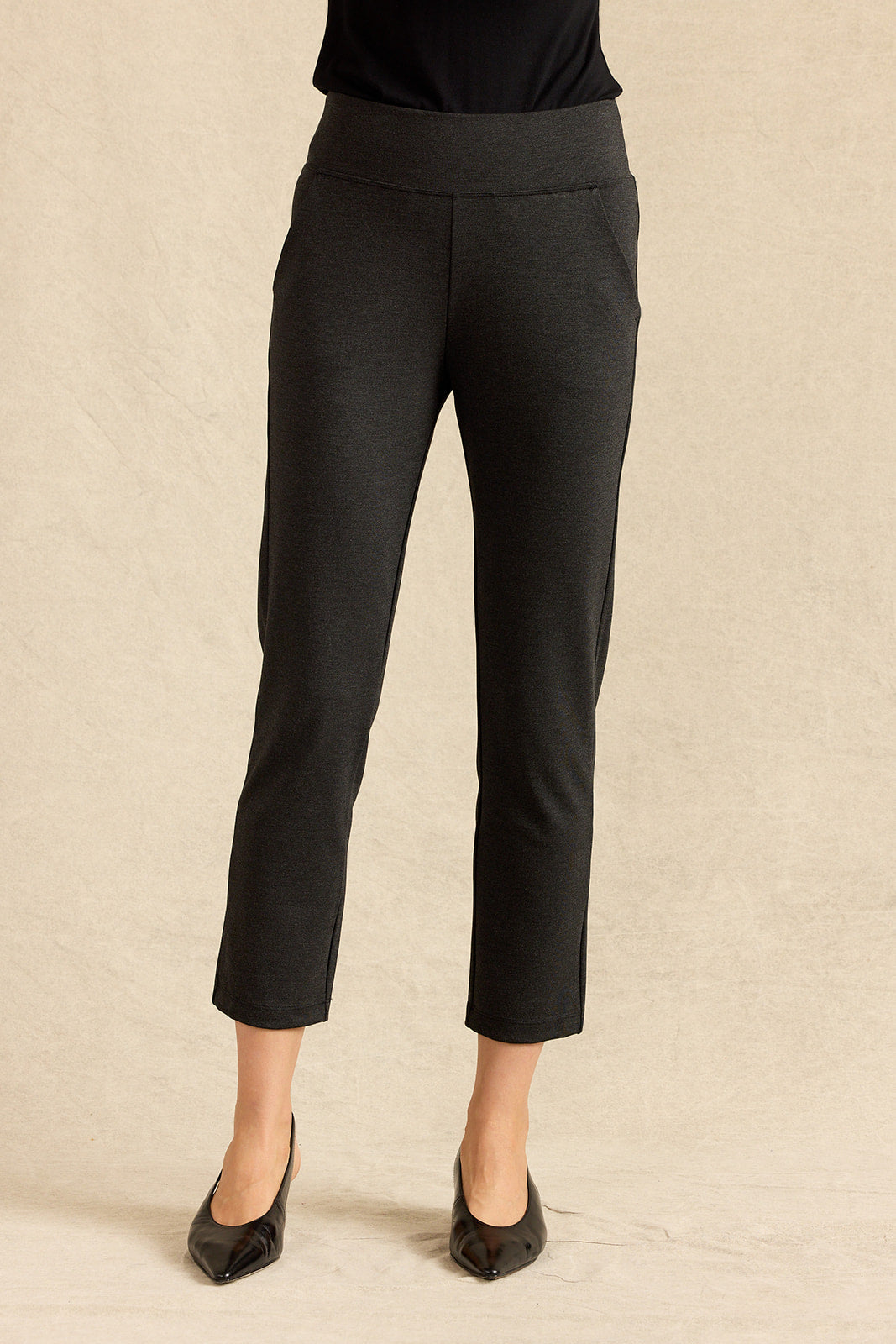 Charcoal cropped ankle-length pants shown on a model from the waist down, paired with a black top and black pointed-toe heels, against a textured beige studio backdrop.