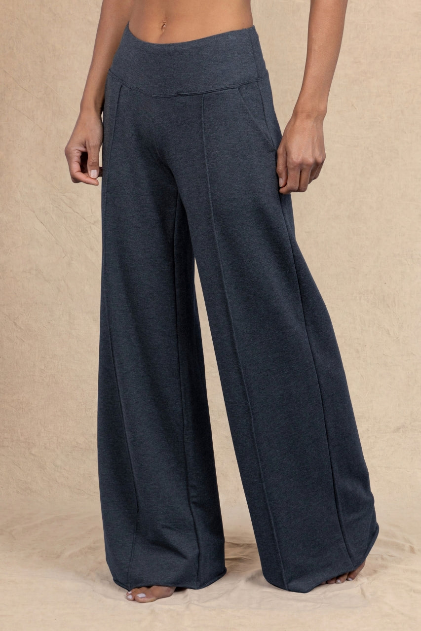 A person wearing dark charcoal wide-leg pants, shown from the waist down against a neutral beige textured backdrop.