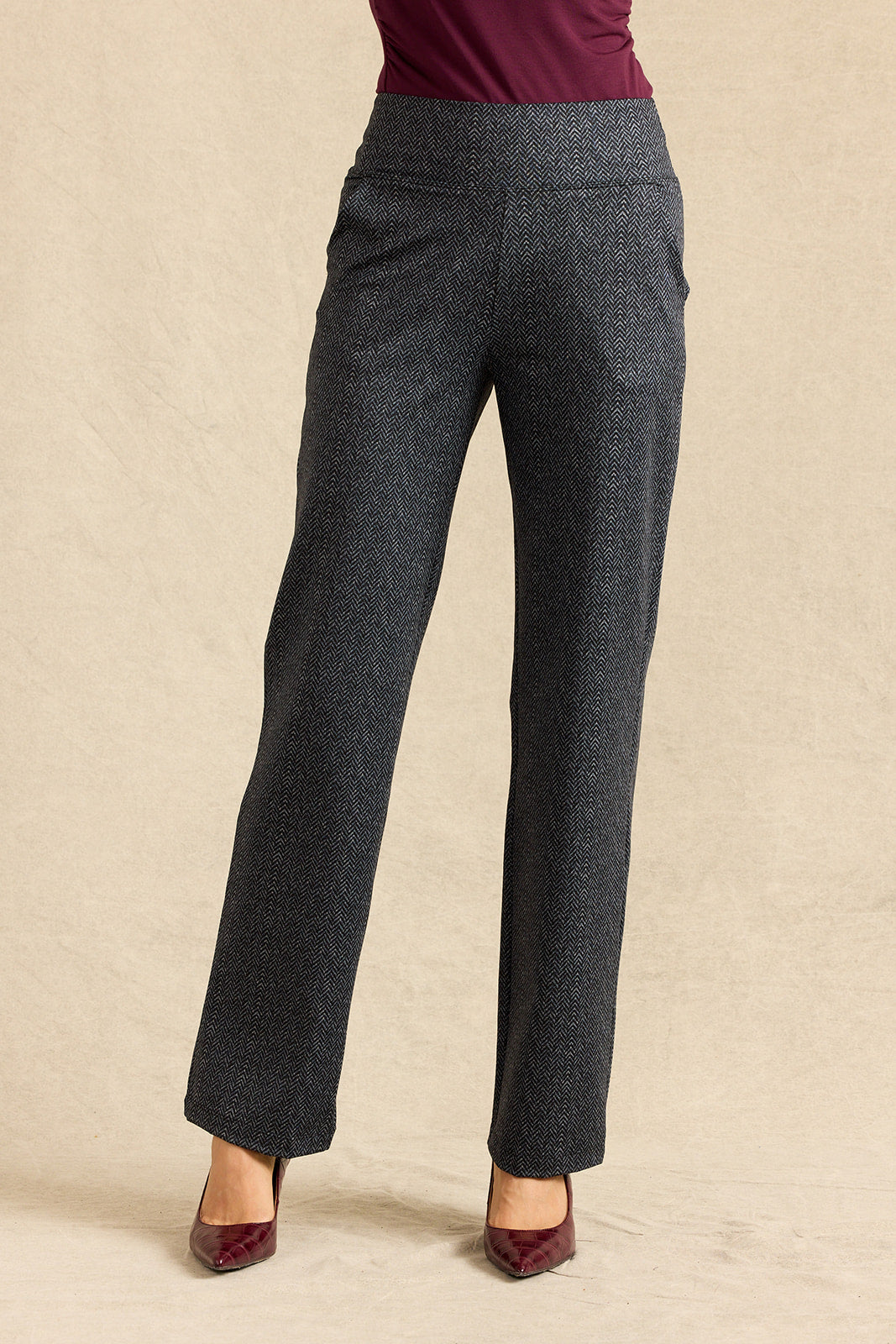 A person wearing dark charcoal gray herringbone trousers, styled with a burgundy top and burgundy pointed-toe heels, standing against a textured beige backdrop.