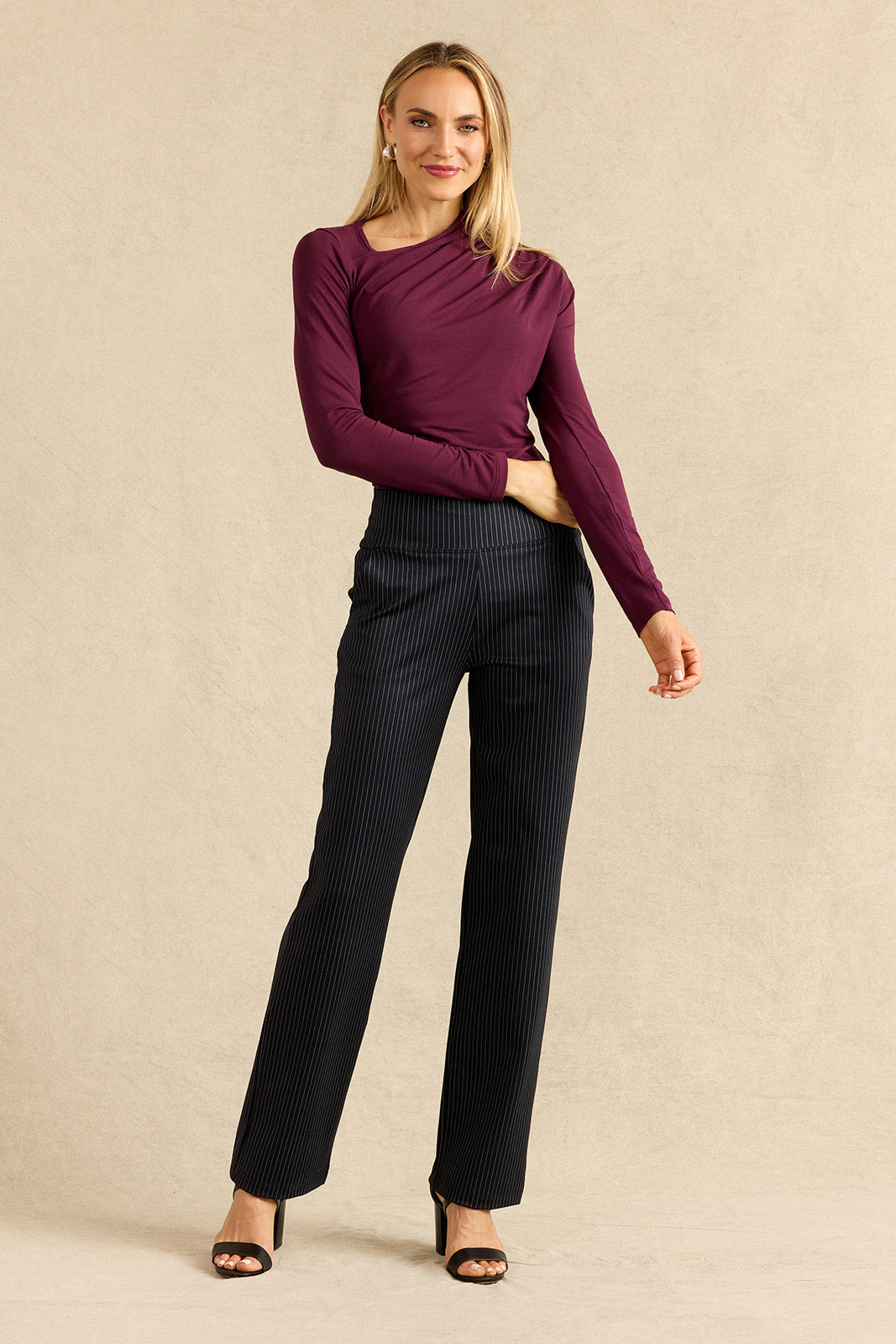 A woman modeling a dark burgundy long-sleeve top and black pinstriped trousers against a textured beige studio backdrop.