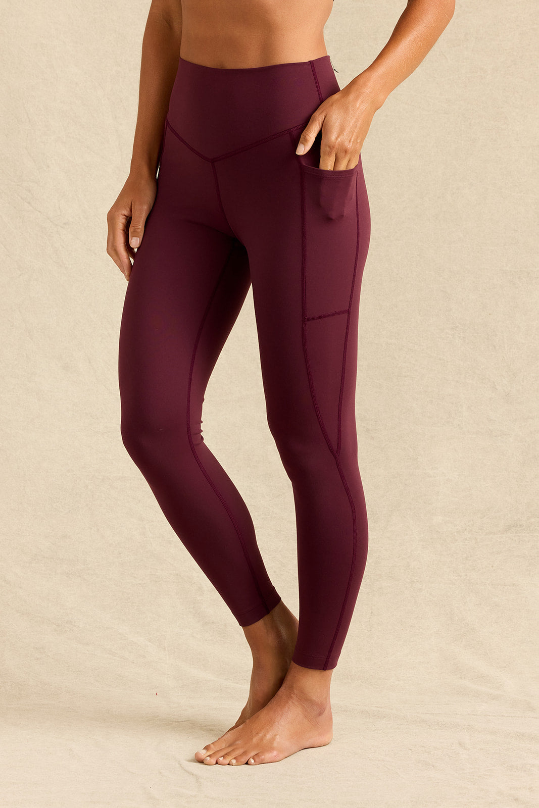 A model wearing high-waisted, ankle-length leggings in a deep burgundy color, featuring a side pocket, standing against a neutral beige backdrop.