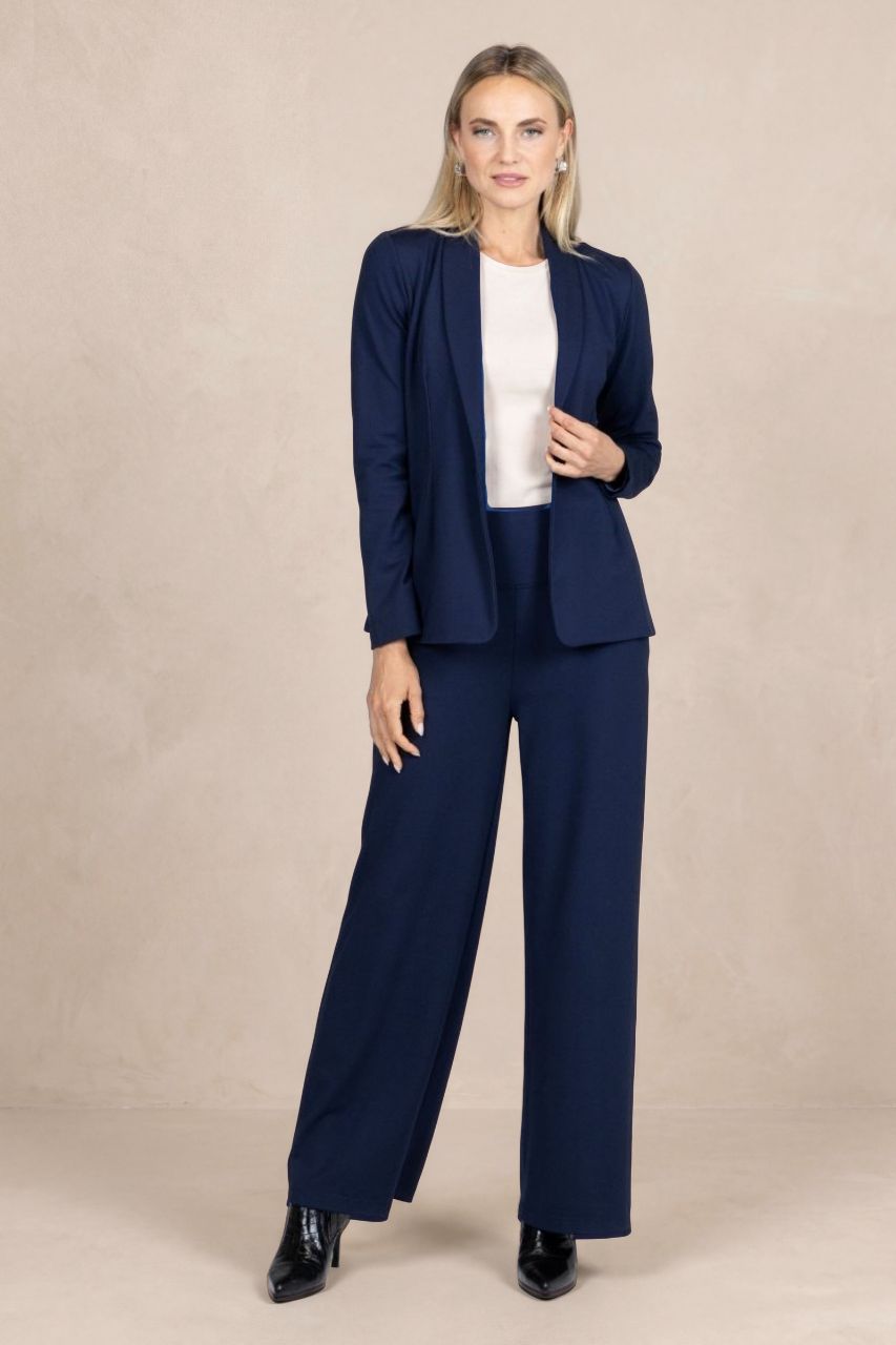 A woman modeling a navy blue two-piece suit consisting of a blazer and wide-leg trousers, styled with a white top and black patent leather shoes, against a neutral beige studio backdrop.