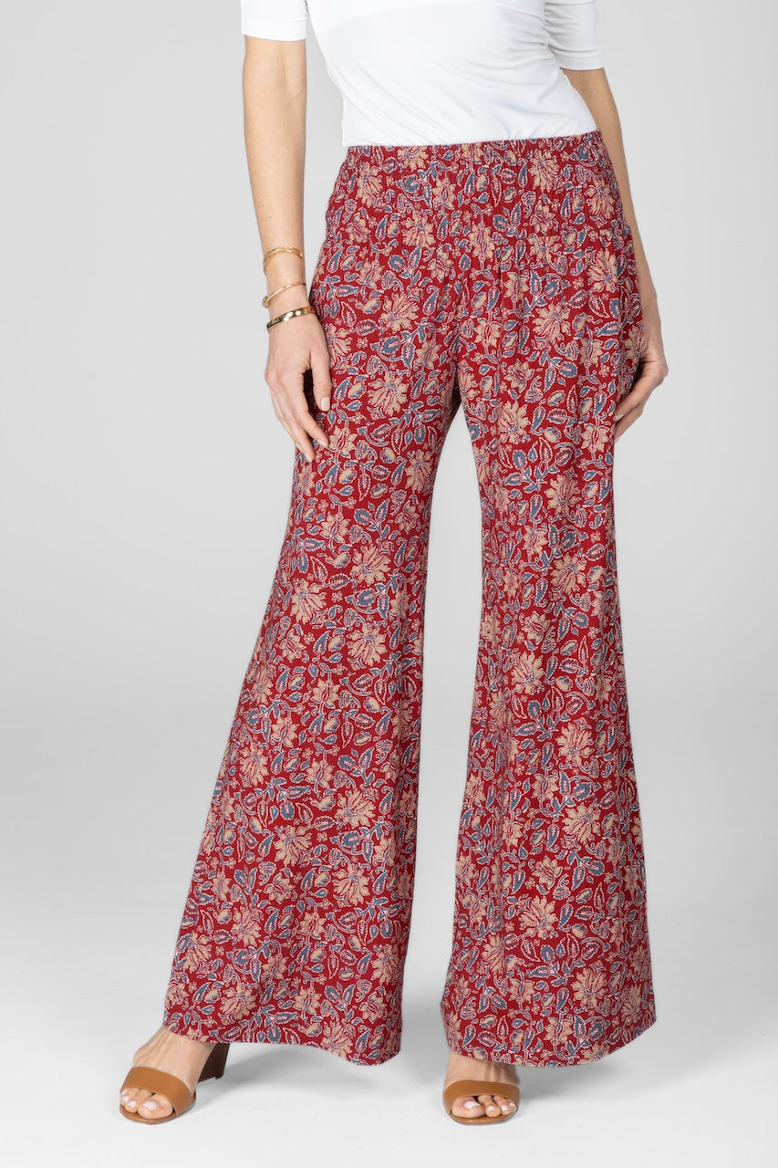 A woman wearing a pair of red motif printed wide-leg pants with a smocked waistband and side pockets, paired with a white short sleeve top and tan sandals.