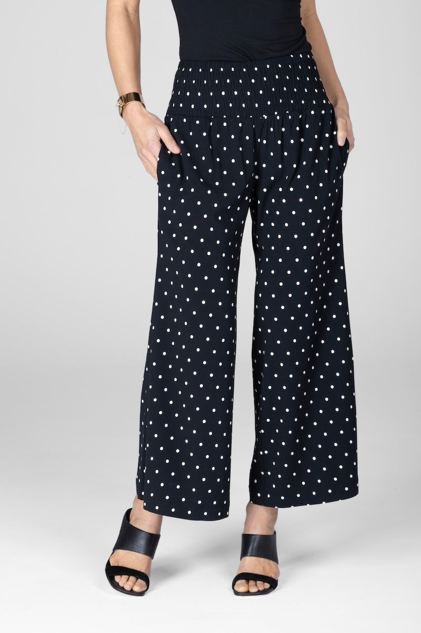 A pair of ankle-length black pants with a white dot print, featuring a smocked waistband and hands in side pockets.