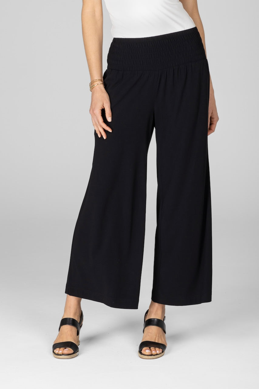 A woman wearing black ankle pants with a smocked waistband and side pockets, paired with a white top and black sandals.