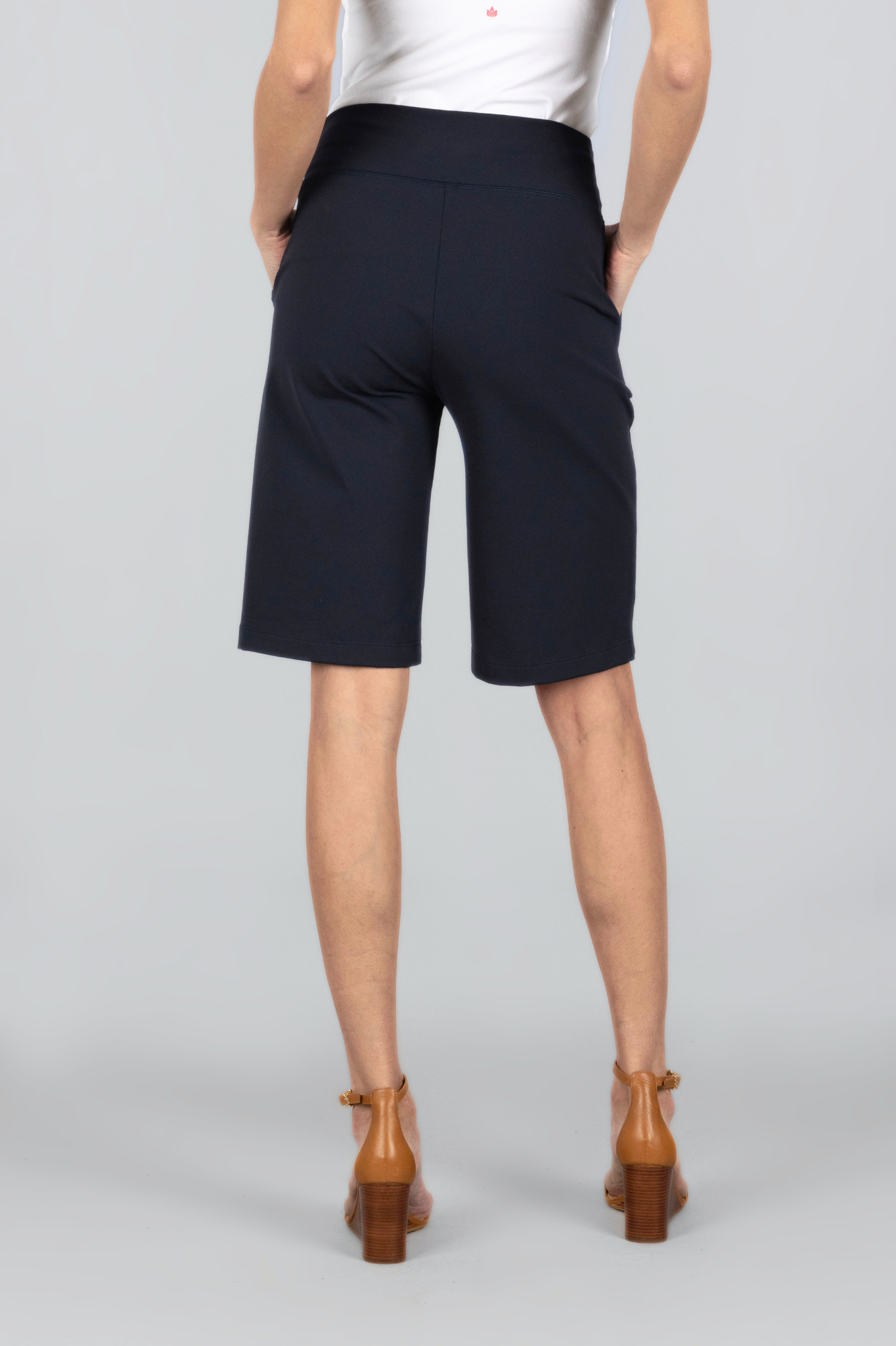 Female model wearing white shirt and black knee length shorts with hands in her pockets. Back view showing the back of shorts