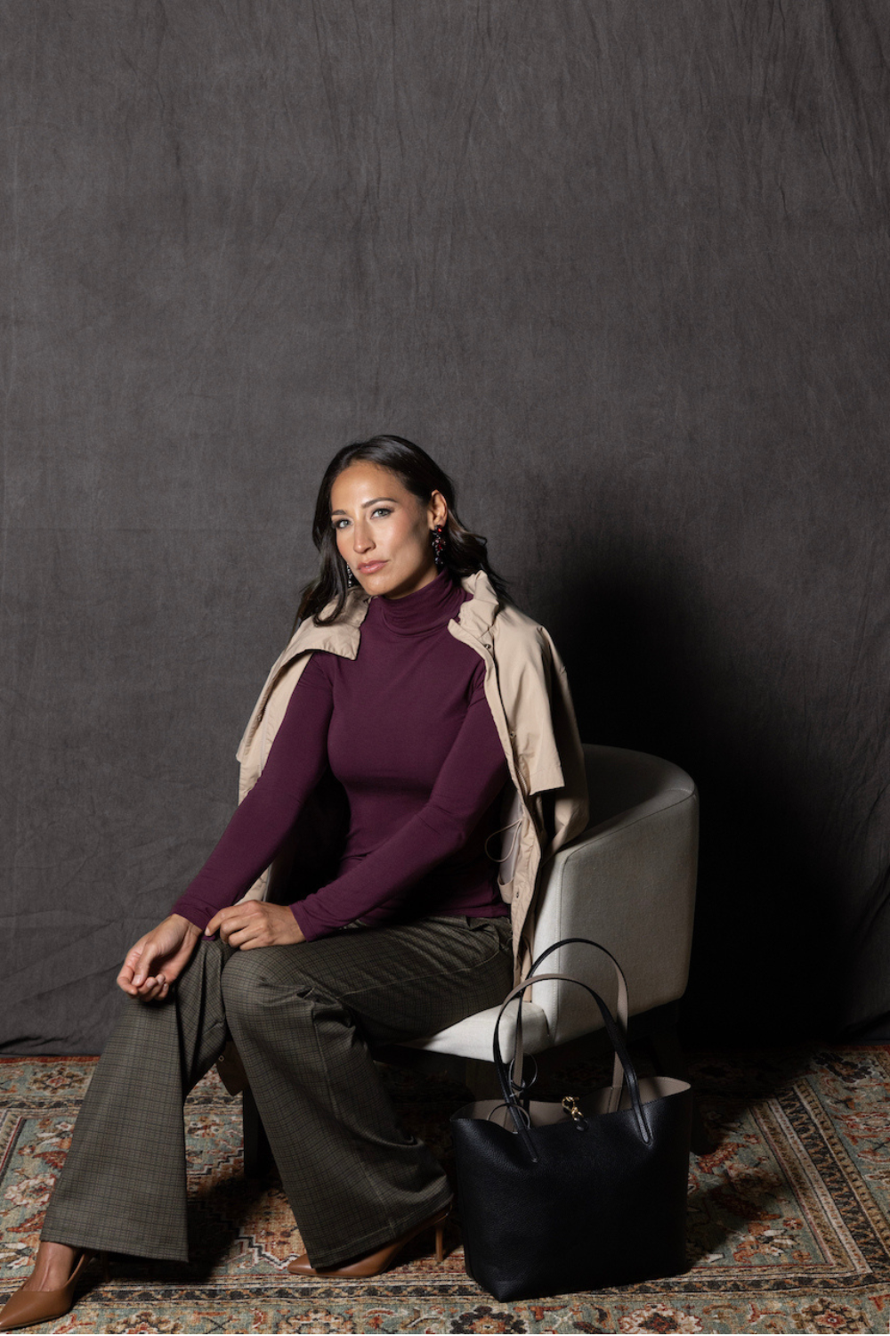 Woman sitting on a chair wearing a burgundy turtleneck top and beige coat draped over her shoulders against a dark background