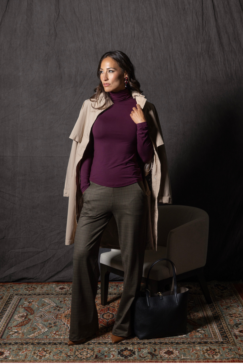Woman in a burgundy turtleneck and beige coat standing against a dark background