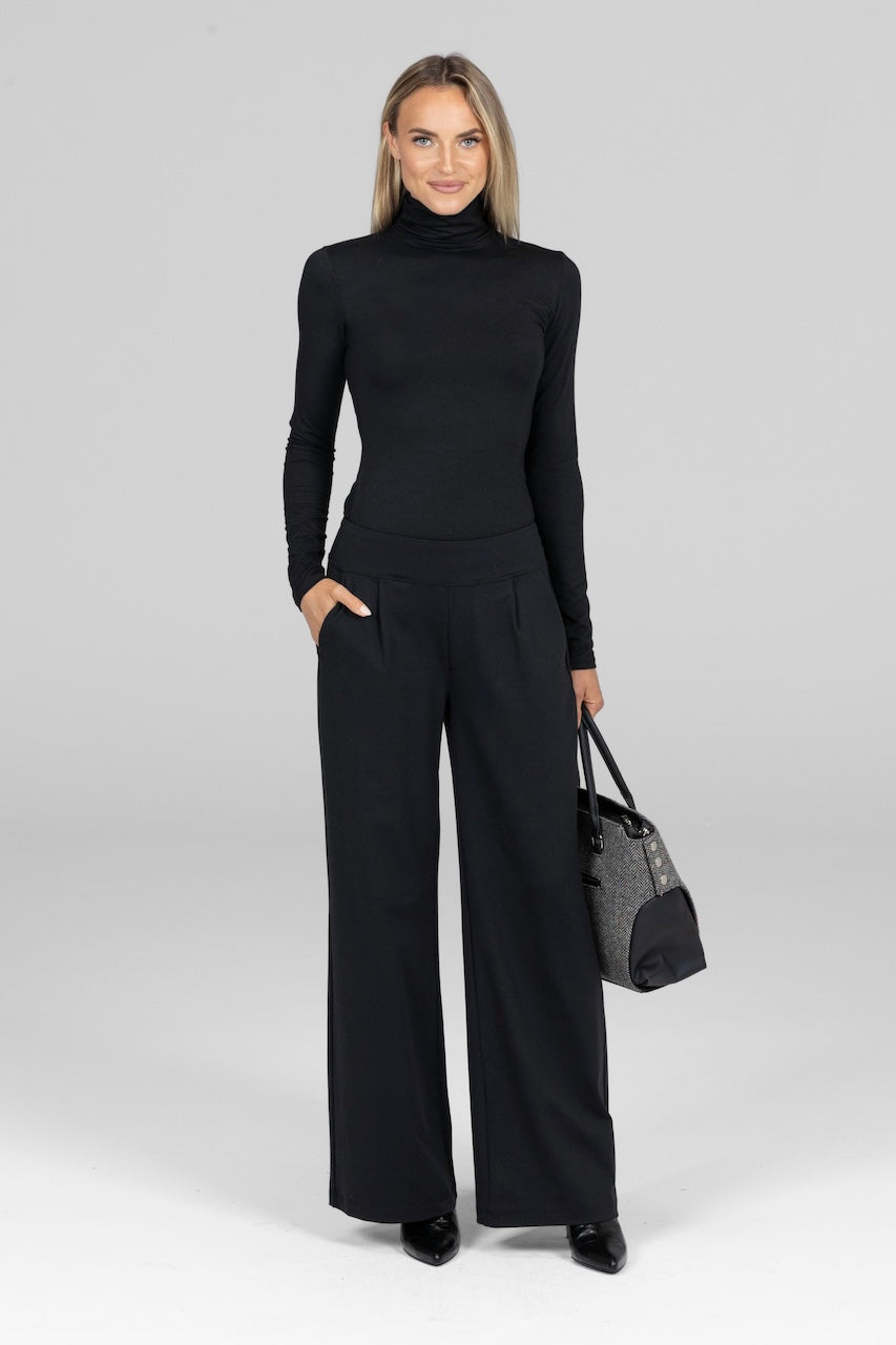 Full length photo of a woman wearing a black turtleneck and black pleated dress pants