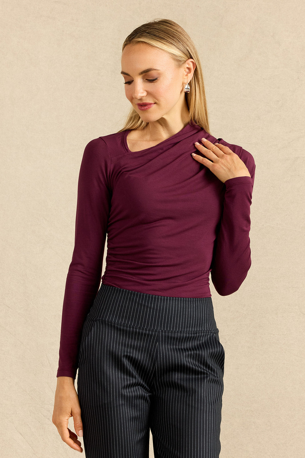 A woman modeling a long-sleeve, asymmetrical-neck top in deep burgundy, paired with dark pinstriped trousers, against a textured beige background.