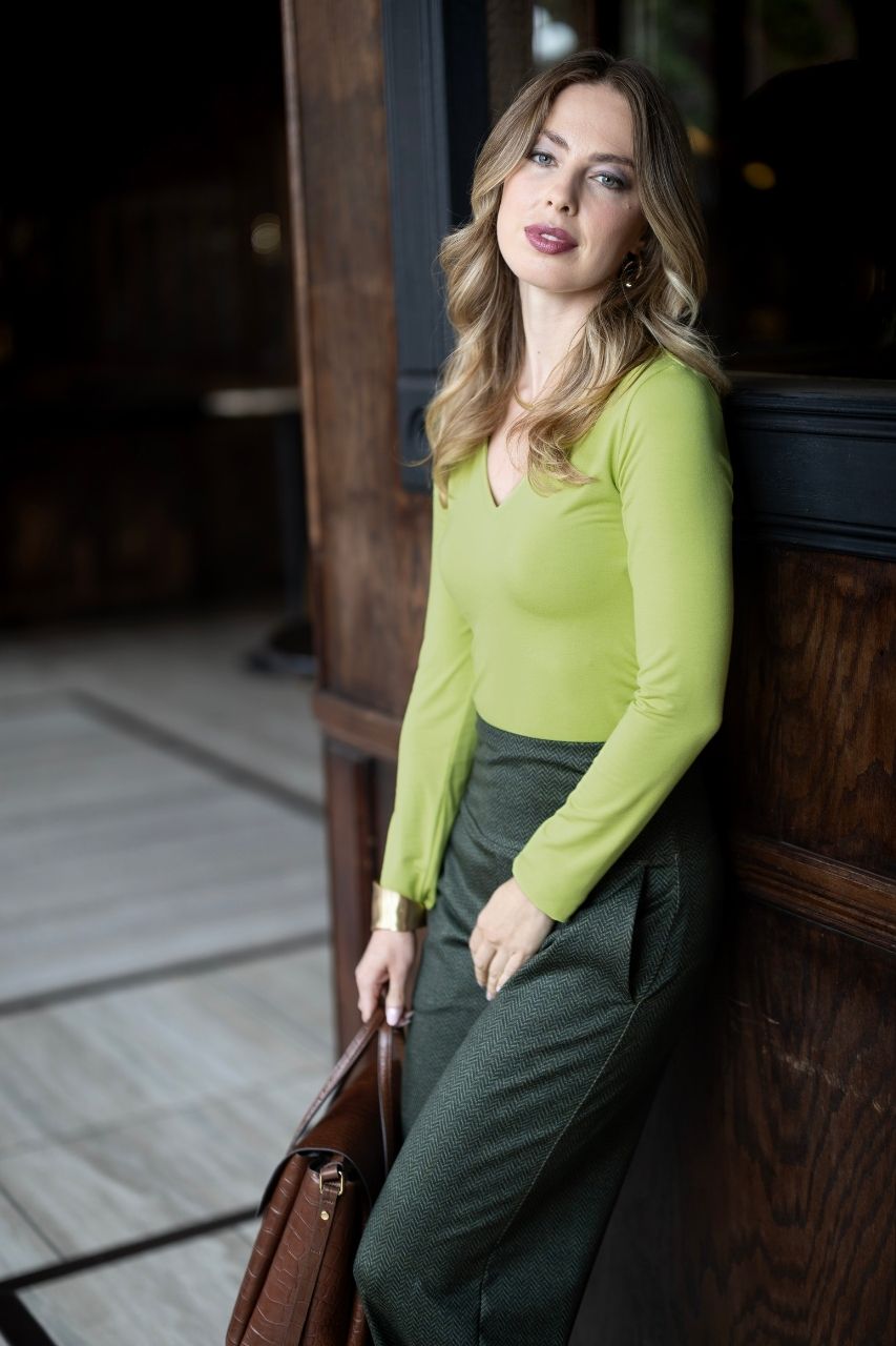 Woman in a chartreuse top and olive tweed high waisted pants leaning against a wooden wall indoors