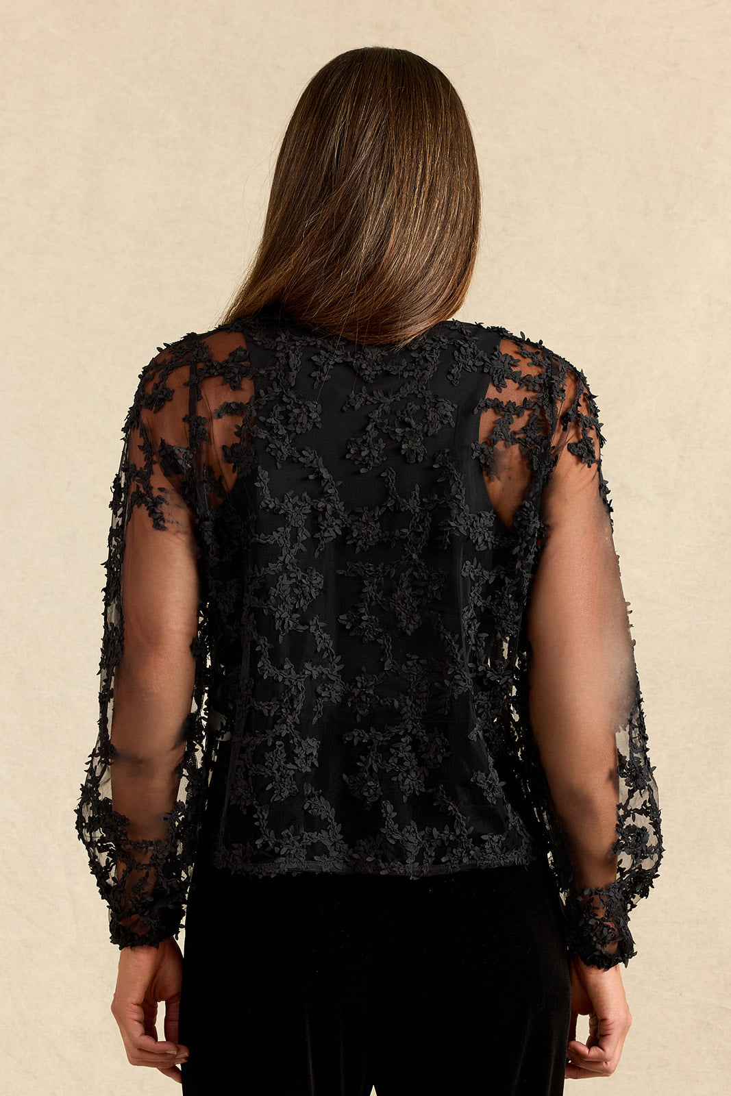 A woman viewed from the back wearing a black lace long-sleeve top with sheer mesh sleeves and black pants, set against a plain beige textured background.