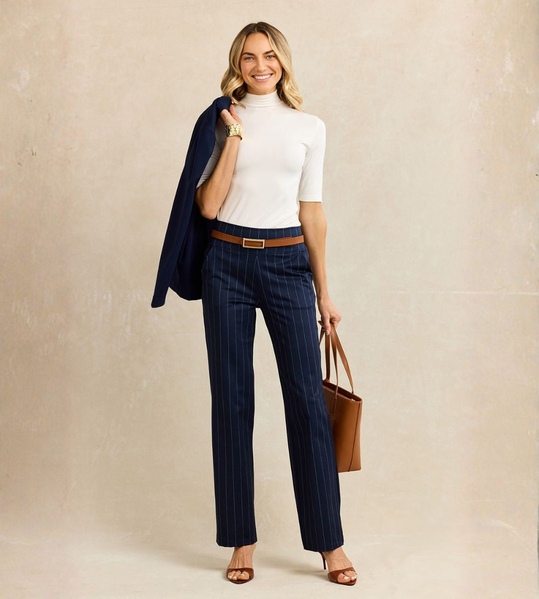 Woman wearing a white mock neck short sleeve top, navy pinstripe pants, and a navy blazer against a beige background