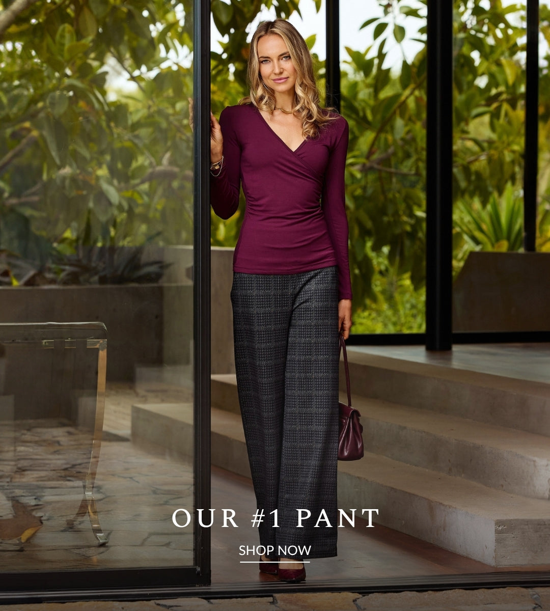 A woman stands in front of large glass doors with lush green foliage outside, wearing a fitted burgundy wrap-style long-sleeve top and dark gray plaid wide-leg pants, holding a matching burgundy handbag. Text at the bottom reads 'OUR #1 PANT SHOP NOW'.