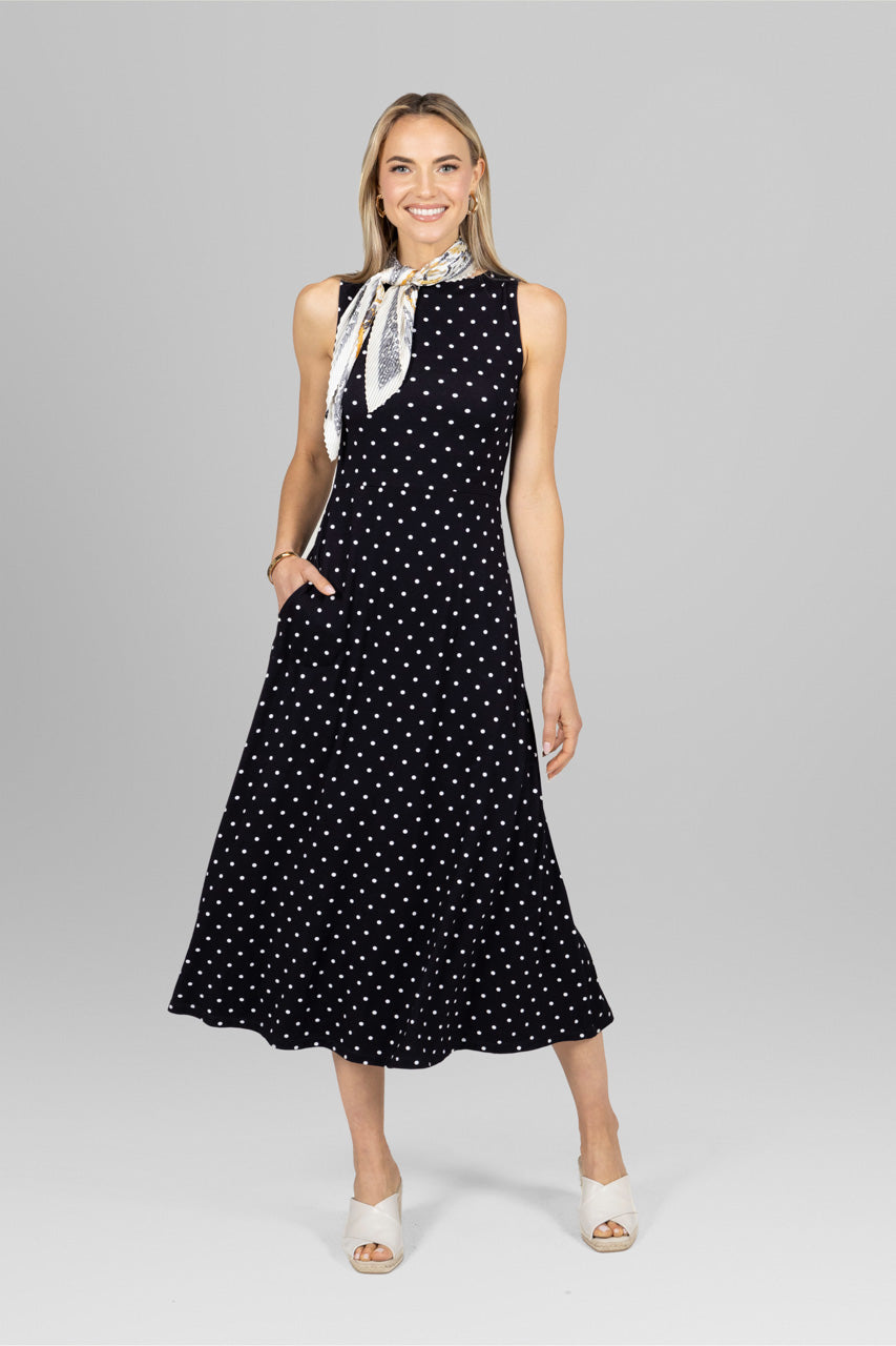 A woman standing in a studio, wearing a sleeveless, black dress with a white dot pattern, a boat neckline, and a flowing A-line silhouette.