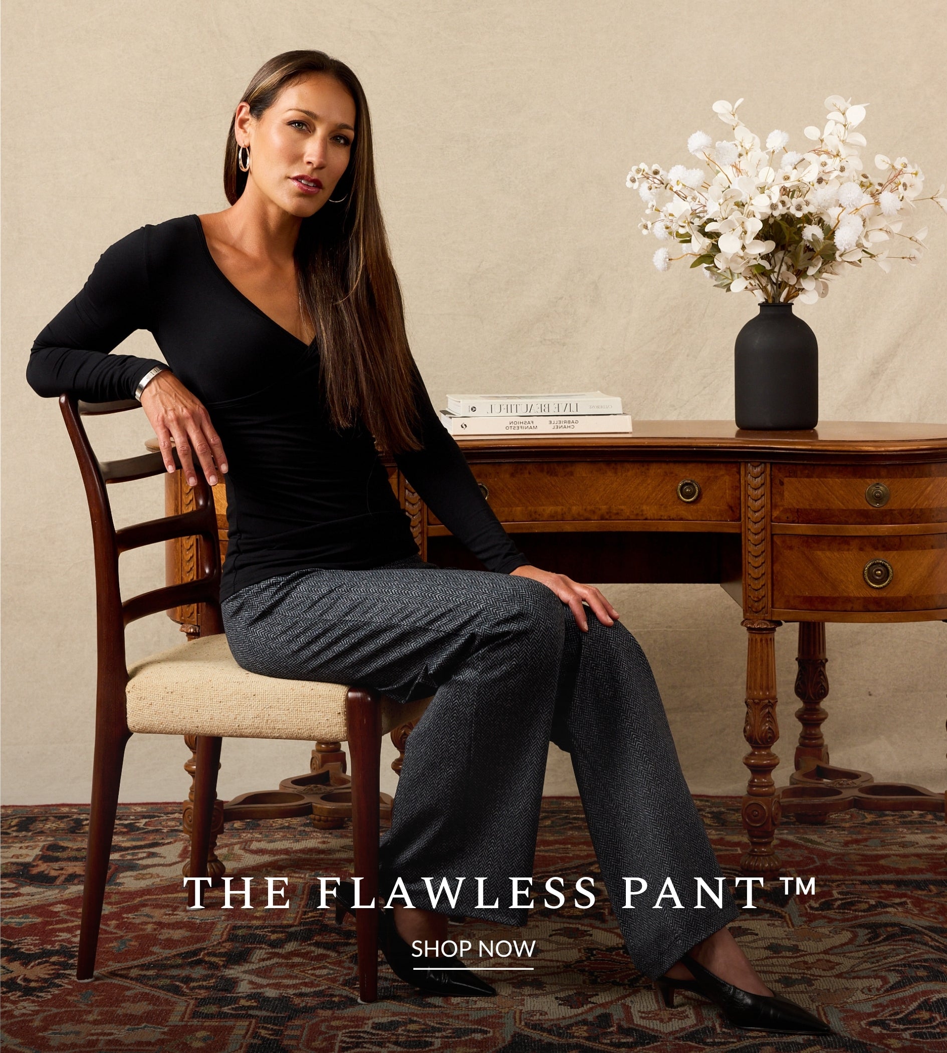 A woman sits on a wooden chair in a styled room, wearing a fitted black long-sleeve V-neck top and dark gray wide-leg pants. A wooden desk with a vase of white flowers is behind her, and text at the bottom reads 'THE FLAWLESS PANT™ SHOP NOW'.