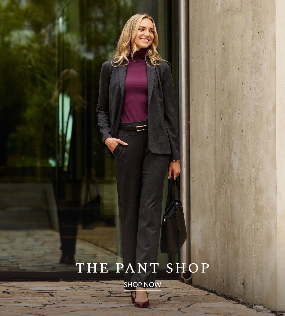 A woman stands outdoors beside a concrete wall and glass storefront, wearing a tailored charcoal blazer and matching straight leg pants over a deep burgundy turtleneck, with a black handbag and heels. Text at the bottom reads 'THE PANT SHOP' and 'SHOP NOW'.