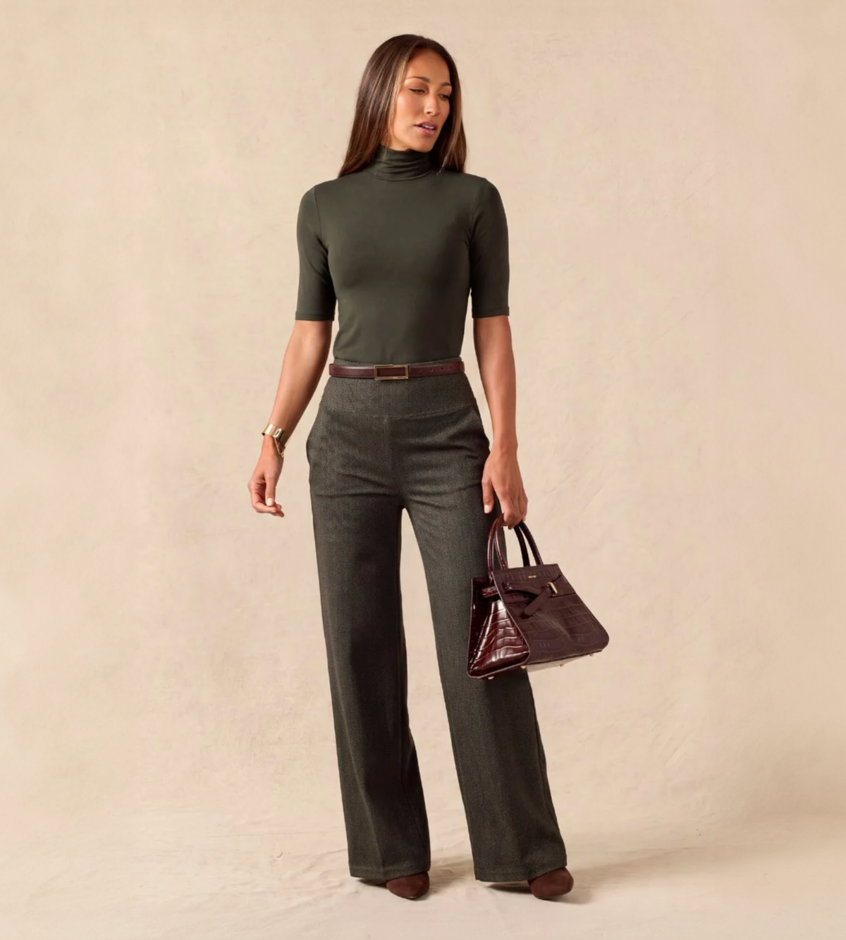 A woman models a dark olive green mock-neck short-sleeve top paired with matching wide-leg trousers, a brown belt, and a dark brown handbag, set against a plain beige studio backdrop.