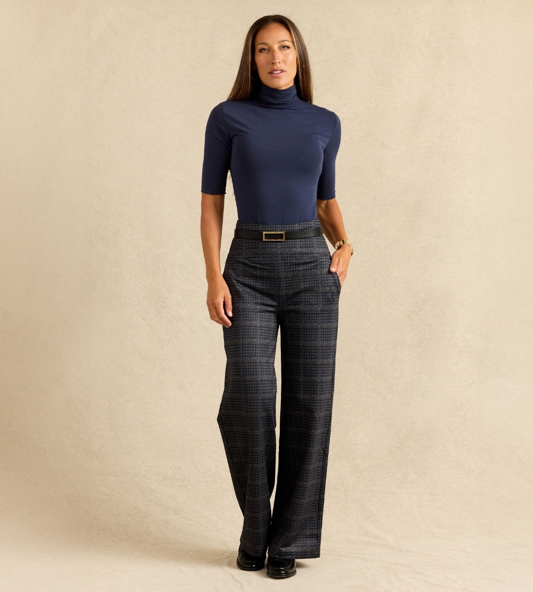 A woman modeling a navy blue short-sleeve turtleneck top paired with dark gray wide-leg plaid trousers, standing against a textured beige studio backdrop.