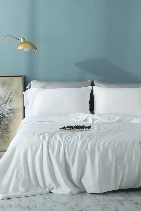White bed sheets on a bed with a blue wall in the background and a gold lamp on the side table.
