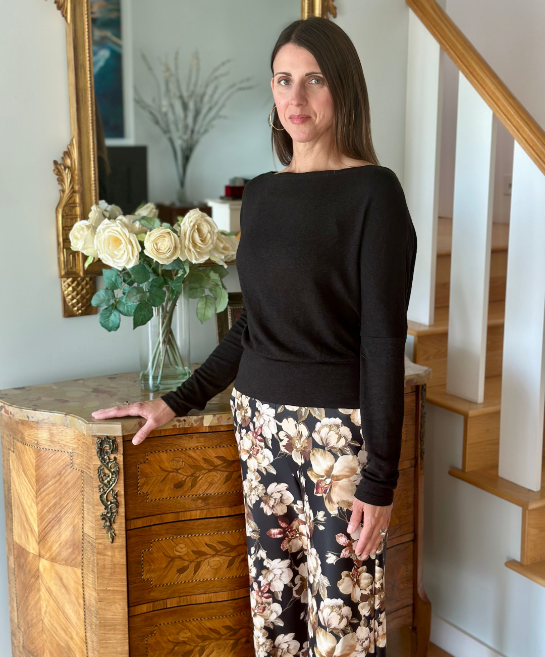 Woman in a black boatneck long sleeve top and floral pants standing in a home setting with a wooden dresser and mirror.