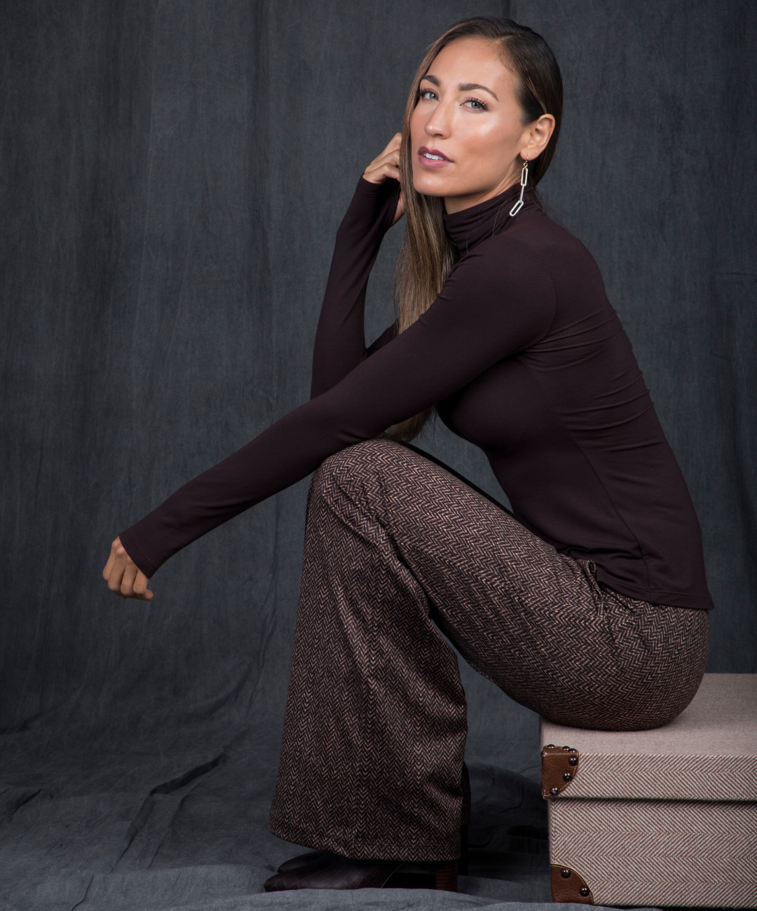 Woman in a dark brown turtleneck and brown herringbone patterned pants sitting against a gray background