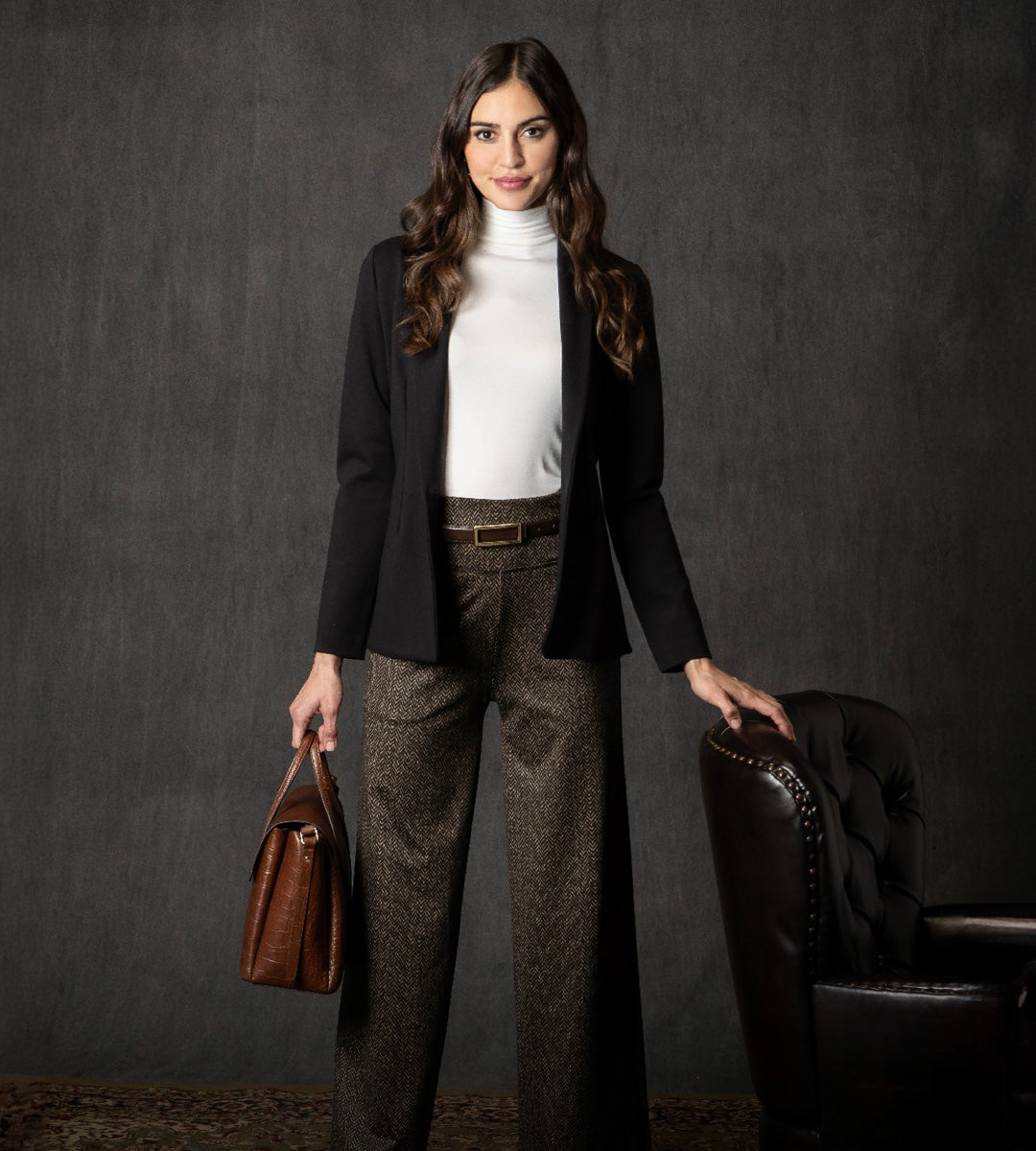 Woman in professional attire with a black blazer and brown herringbone pants, holding a brown leather bag against a dark background.