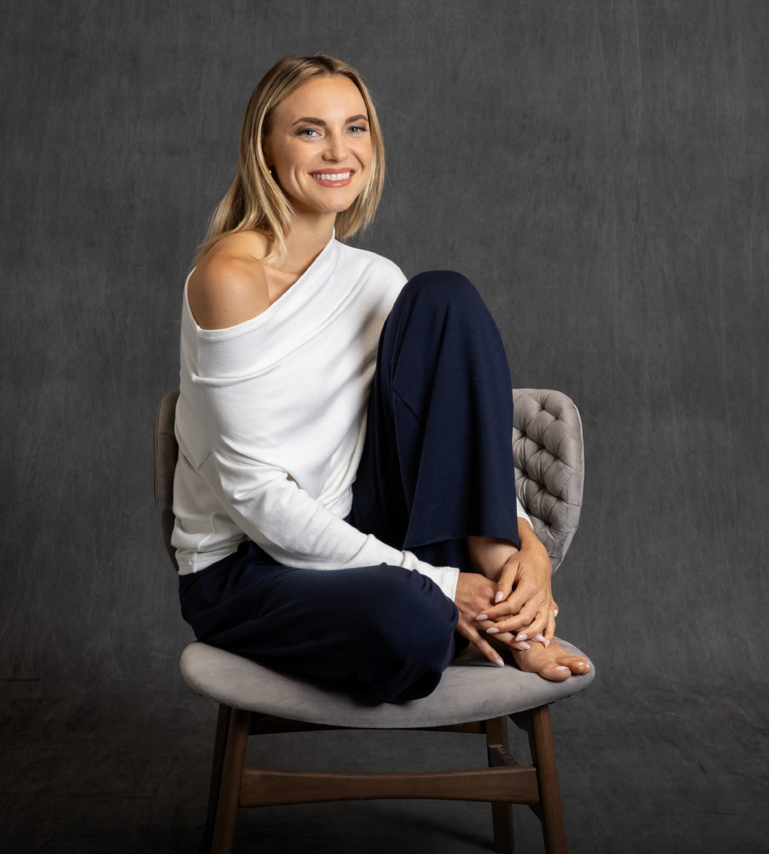 Woman wearing a white off the shoulder sweater and navy blue lounge pants sitting on a chair against a dark gray background