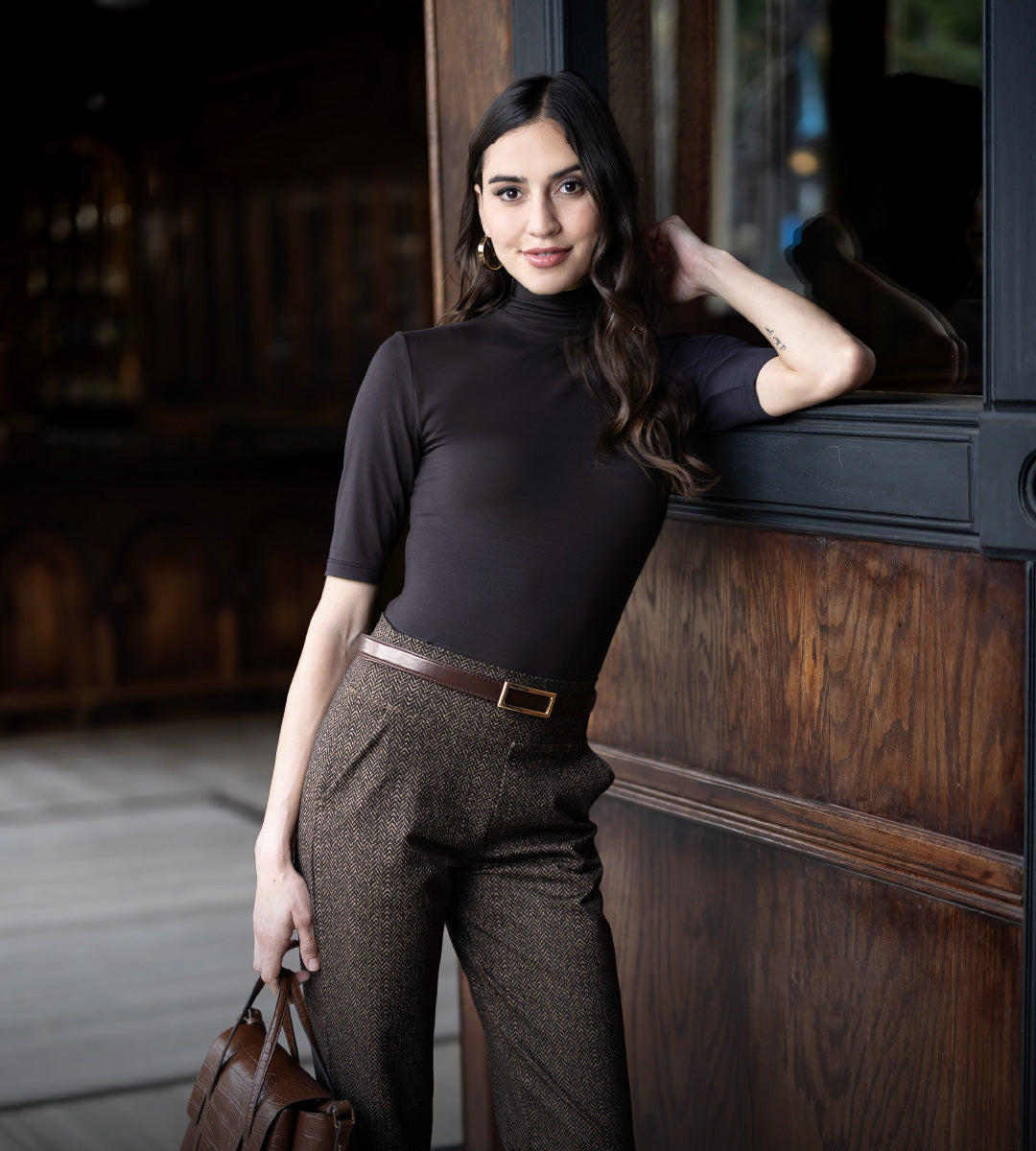 Woman in brown mockneck short sleeve top and brown herringbone patterned pants leaning against a wooden wall.