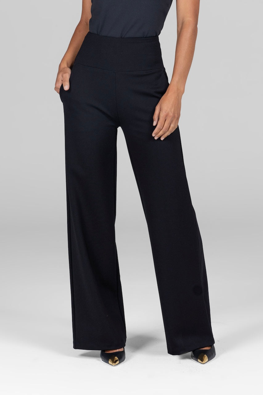 A woman is standing with one hand in her pocket, wearing black high-waist, wide-leg ponte pants. The waistband is thick and structured and the fabric appears soft and slightly stretchy.