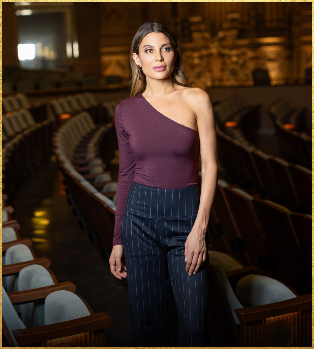 Woman in a one-shoulder burgundy top and pinstripe pants standing in an empty theater.