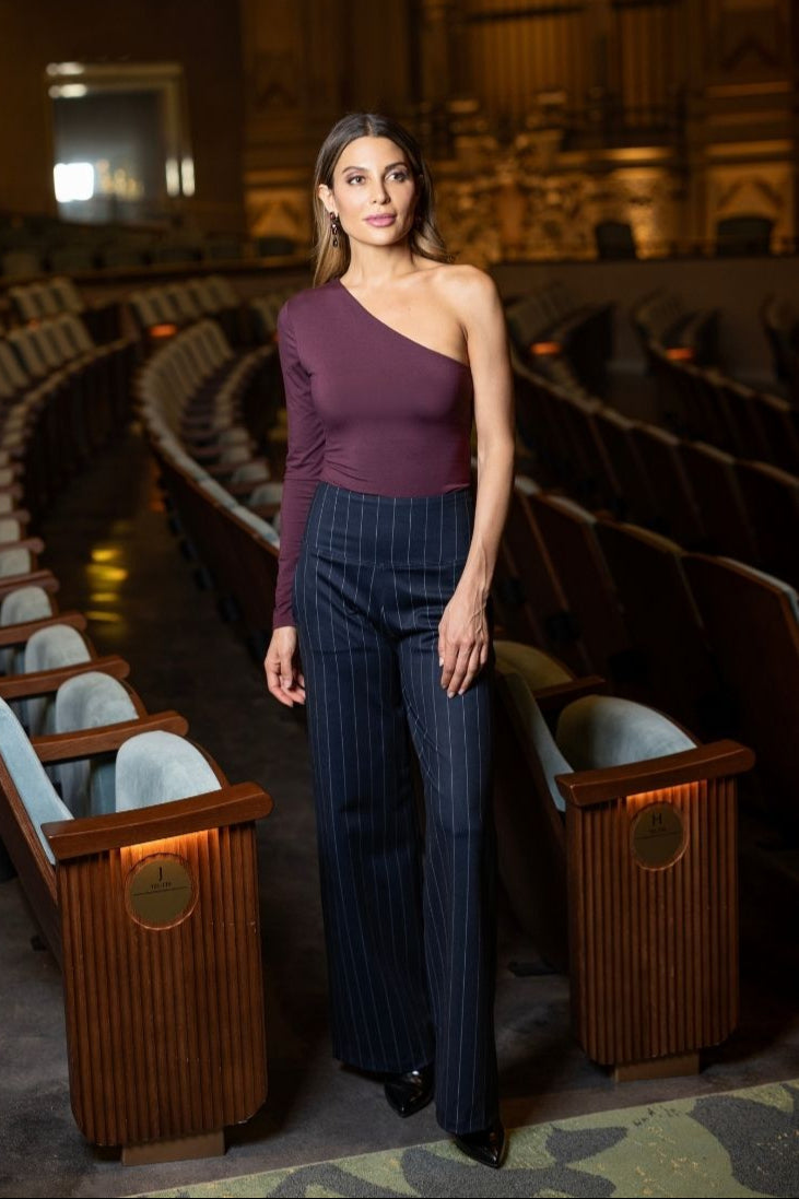 Woman in a burgundy one sleeve top and pinstripe wide leg pants standing in an empty theater.