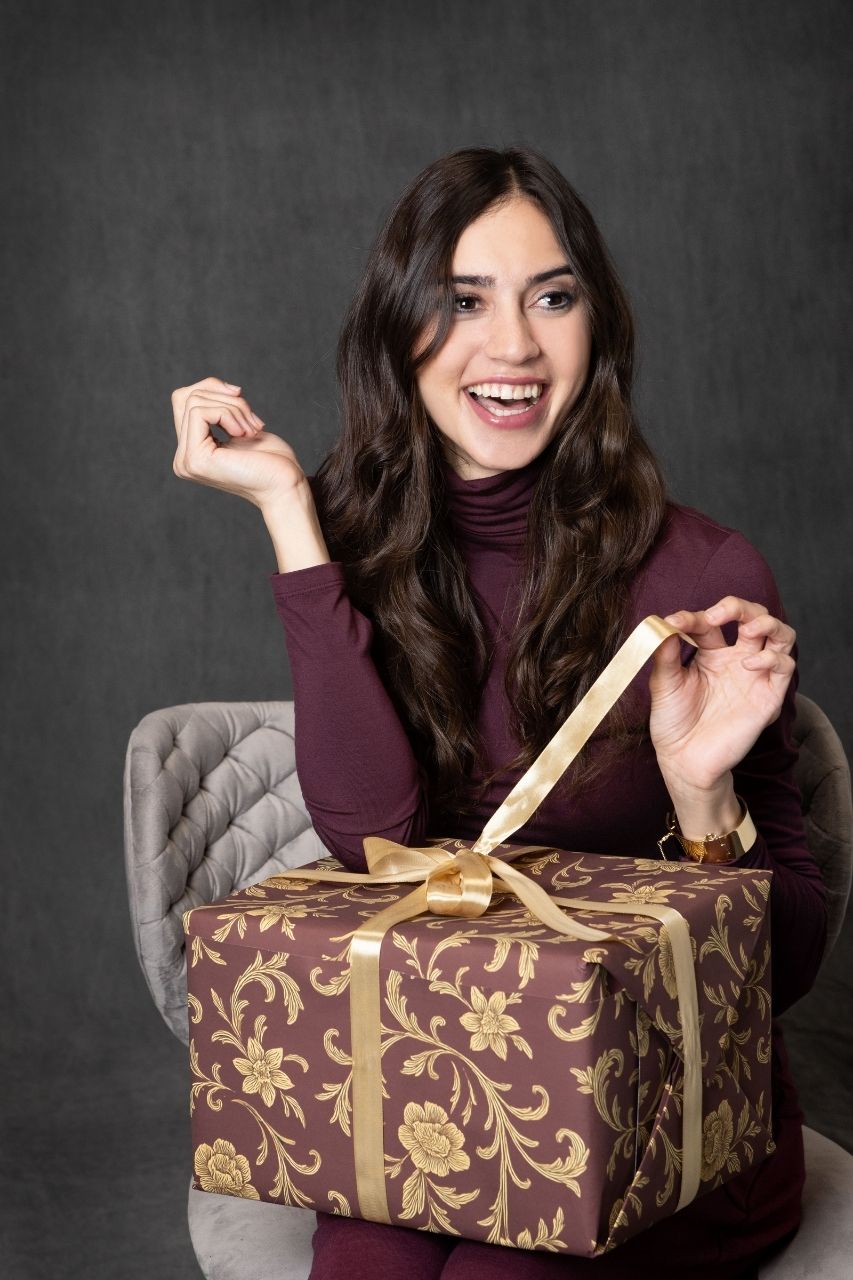 Woman sitting on a chair wearing a burgundy sleeveless turtleneck unwrapping a burgundy and gold gift box