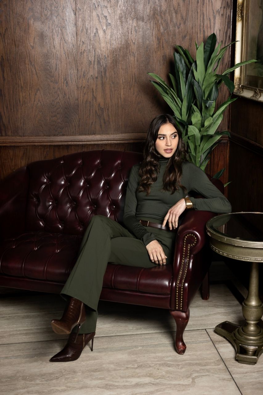 Woman in a green long sleeve turtleneck and matching flared pants sitting on a leather couch in a room with wooden walls and a plant.