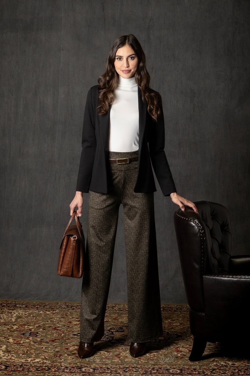 Woman wearing a black blazer, white mockneck tee, and brown printed wide leg pants, holding a brown leather bag against a dark background