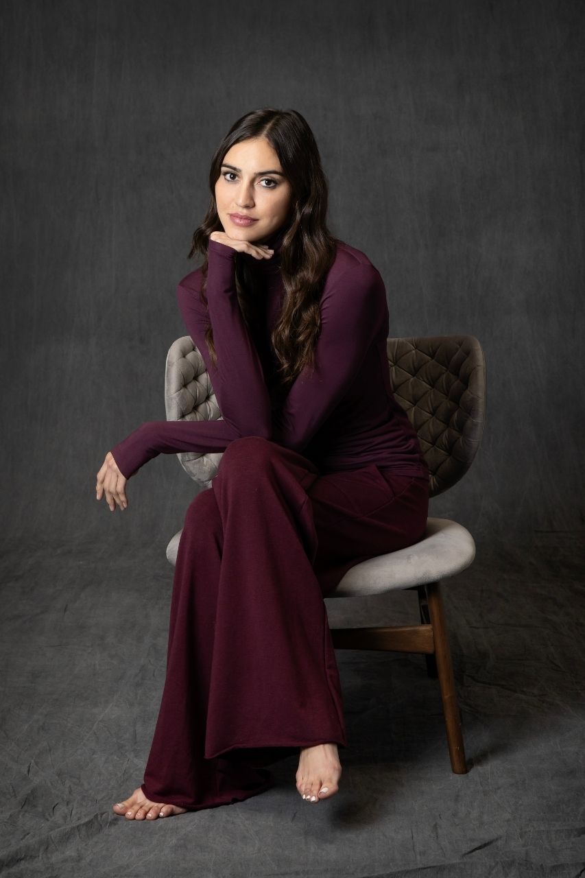 Woman in a burgundy cozy wide leg pant with burgundy turtleneck sitting on a chair against a dark background