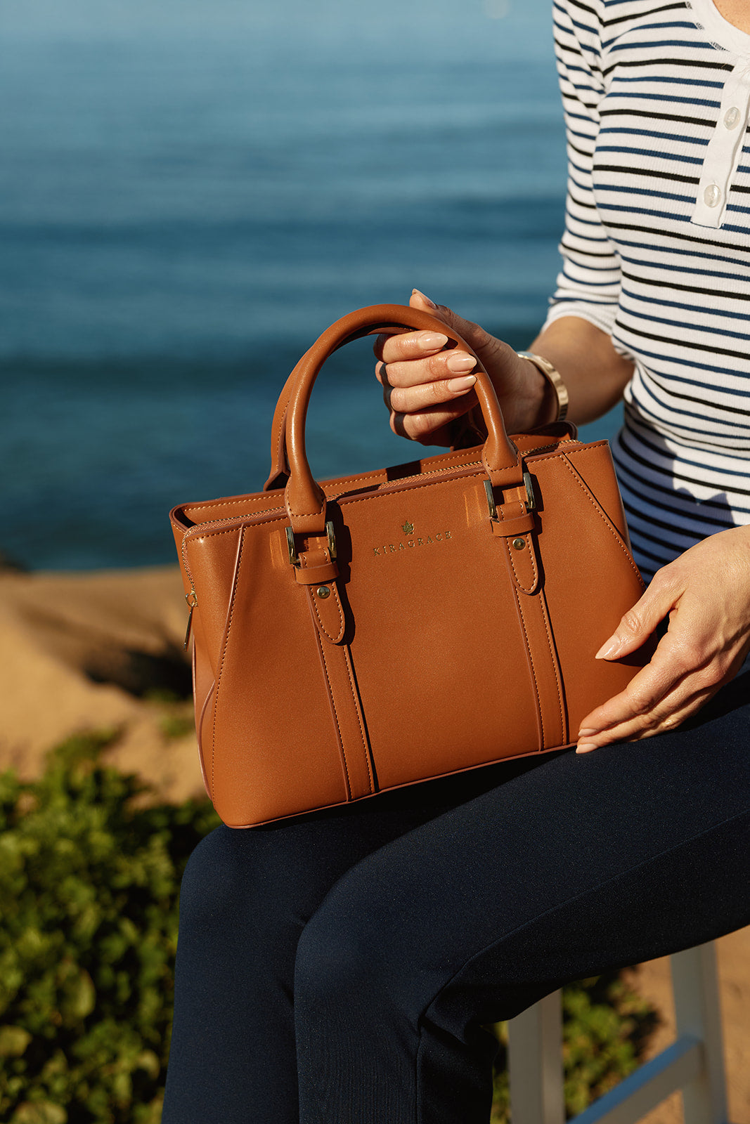 Person holding a brown KiraGrace leather handbag with a blurred ocean background