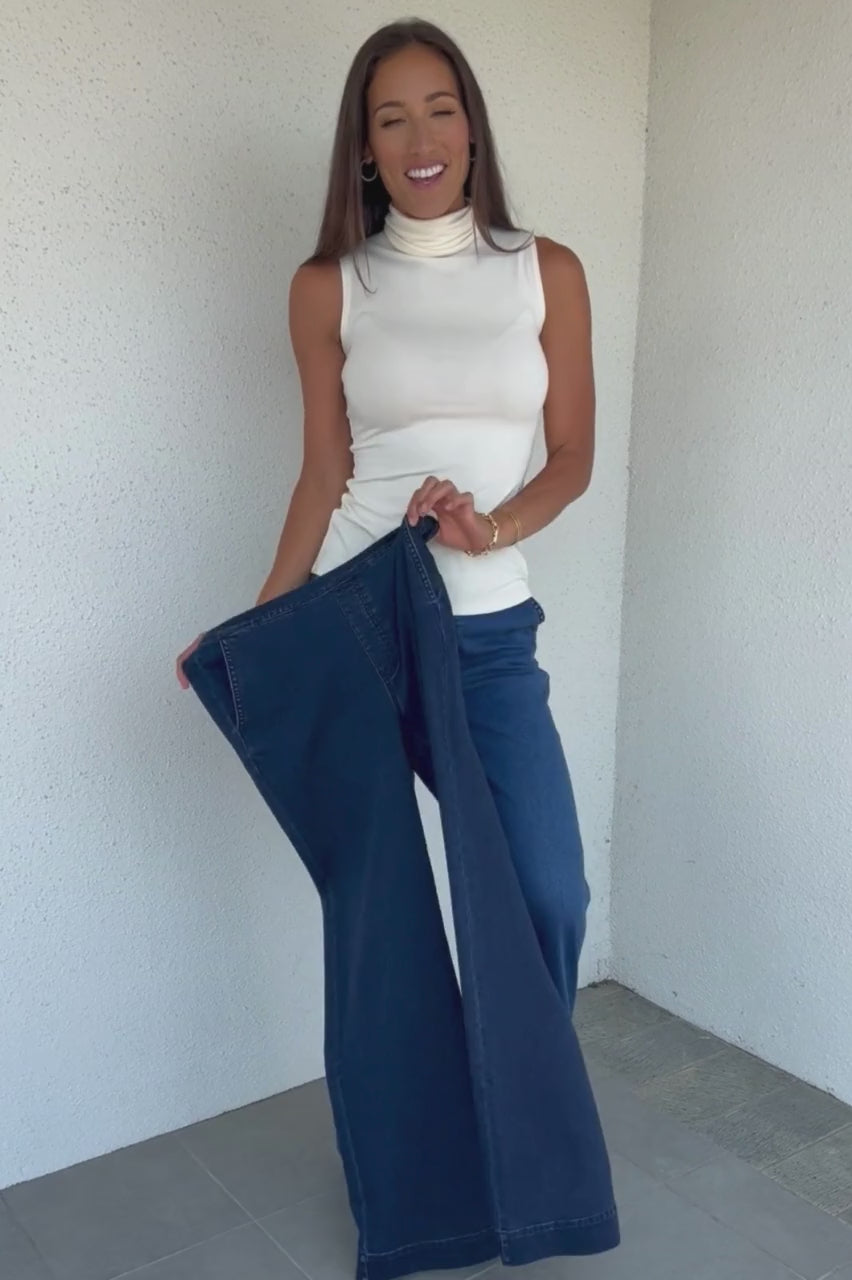 Video of woman wearing white sleeveless turtleneck and light wash stretch denim trousers holding up and stretching a dark wash pair of denim trousers as well as squatting
