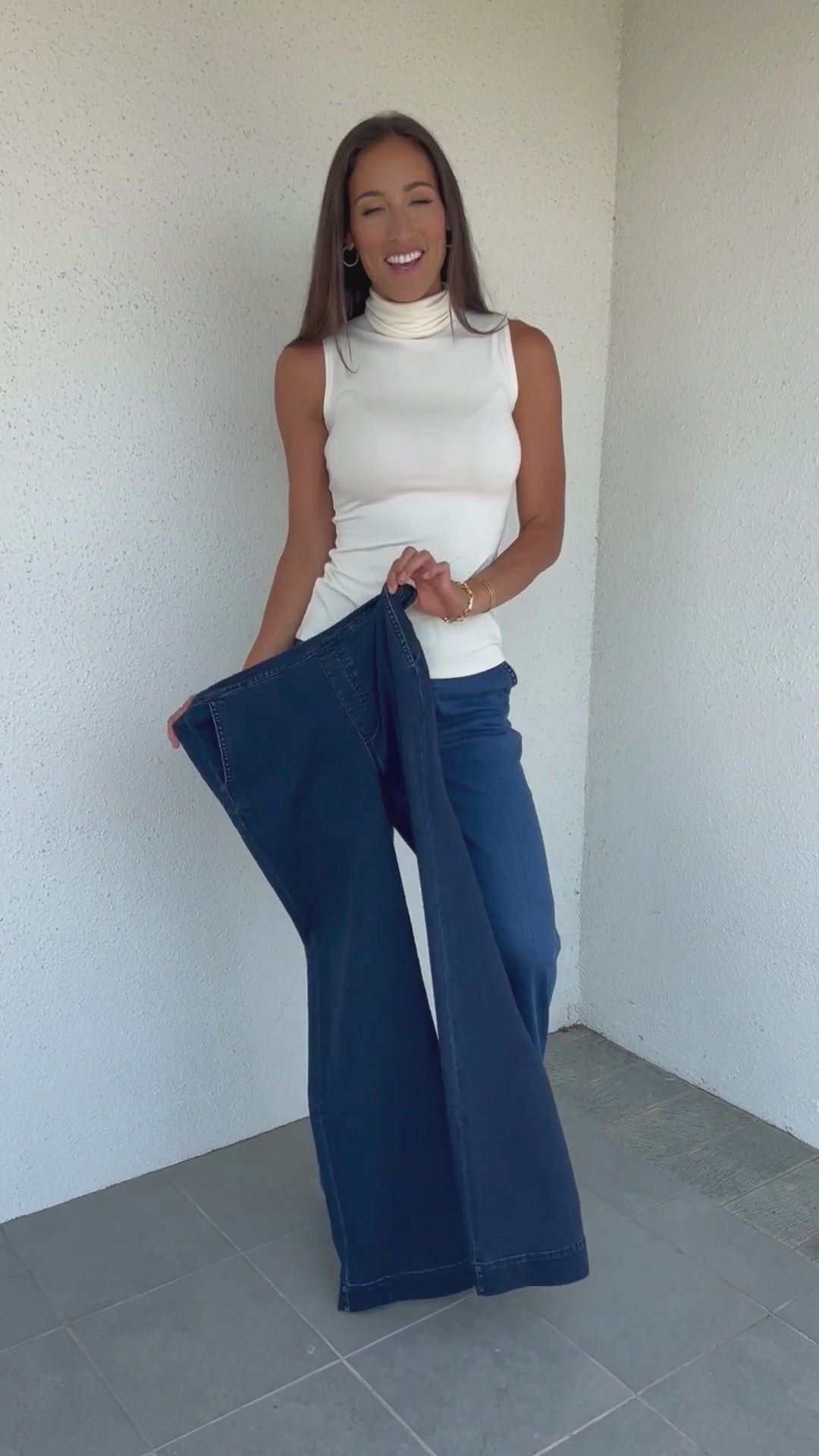 Video of woman holding a pair of dark wash stretch denim trousers showing the stretch, and squatting in the light wash denim trousers