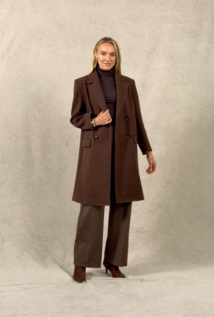 Woman walking onto a studio background wearing a brown coat, brown herringbone pants, and brown boots. She poses for a second, spins, and walks off
