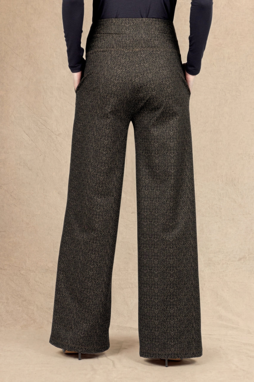 A person shown from the back wearing high-waisted, wide-leg trousers in a dark brown and black textured herringbone pattern, paired with a dark brown long-sleeve top, against a neutral beige backdrop.