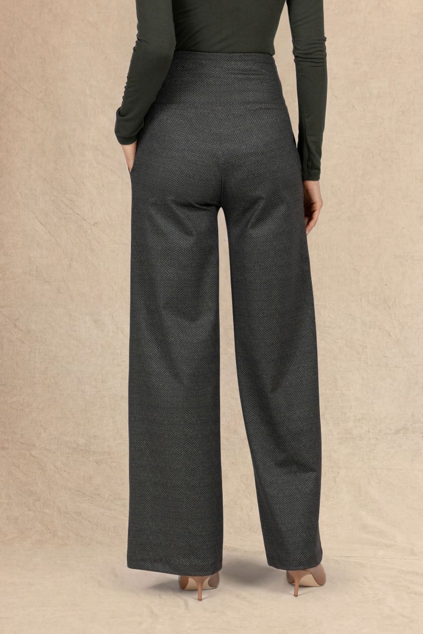A model shown from the back wearing dark olive tweed wide-leg trousers and a dark loden green long-sleeve top, standing against a textured beige backdrop.