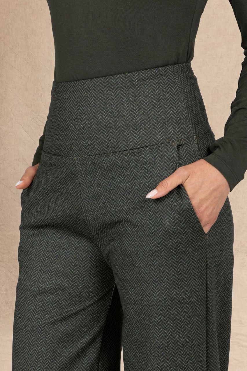 Close-up of a person wearing dark olive tweed herringbone patterned trousers with side pockets, paired with a black long-sleeve top, against a neutral beige background.