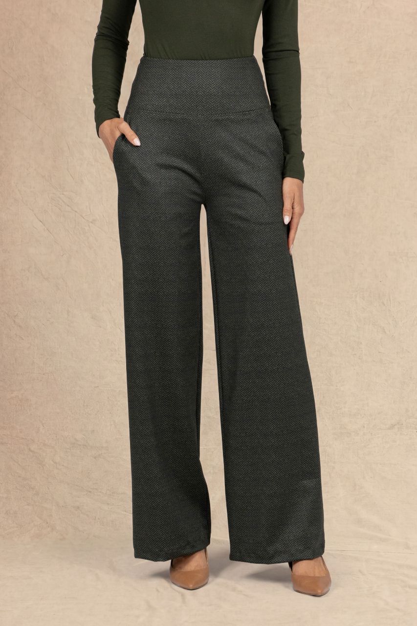 A model wearing dark olive tweed wide-leg trousers with side pockets, paired with an loden green long-sleeve top and tan pointed-toe heels, standing against a textured beige backdrop.